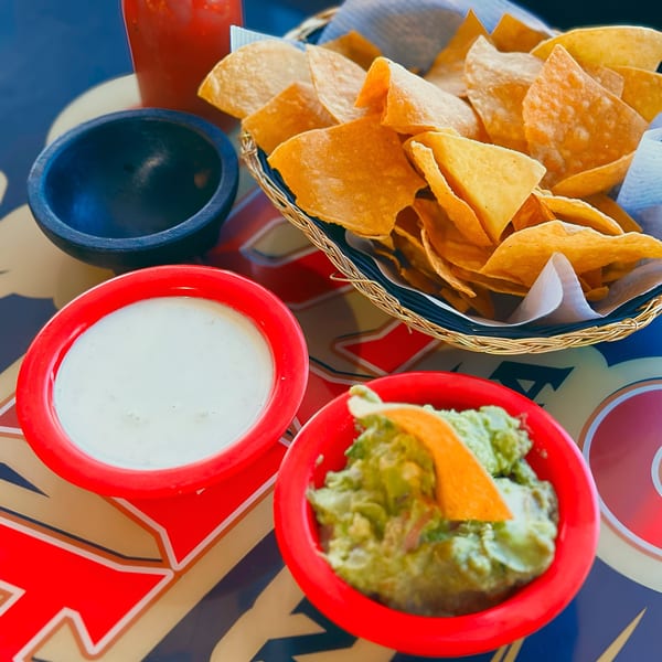 A basket of tortilla chips with bowls of guacamole and queso dip on a colorful table.